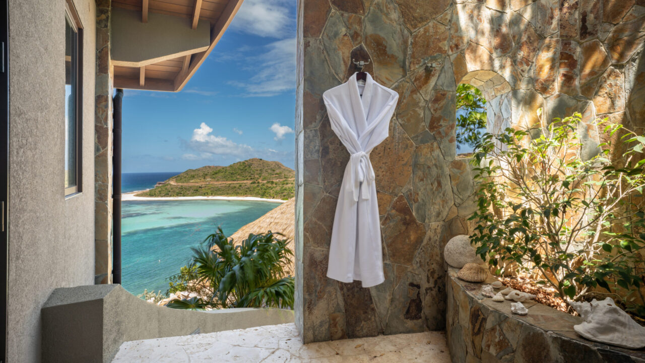 An open-air luxury villa shower alcove at Oil Nut Bay featuring natural local stone walls, a plush white robe, and a stunning arched window view looking out over the turquoise waters and green hillsides of Virgin Gorda.