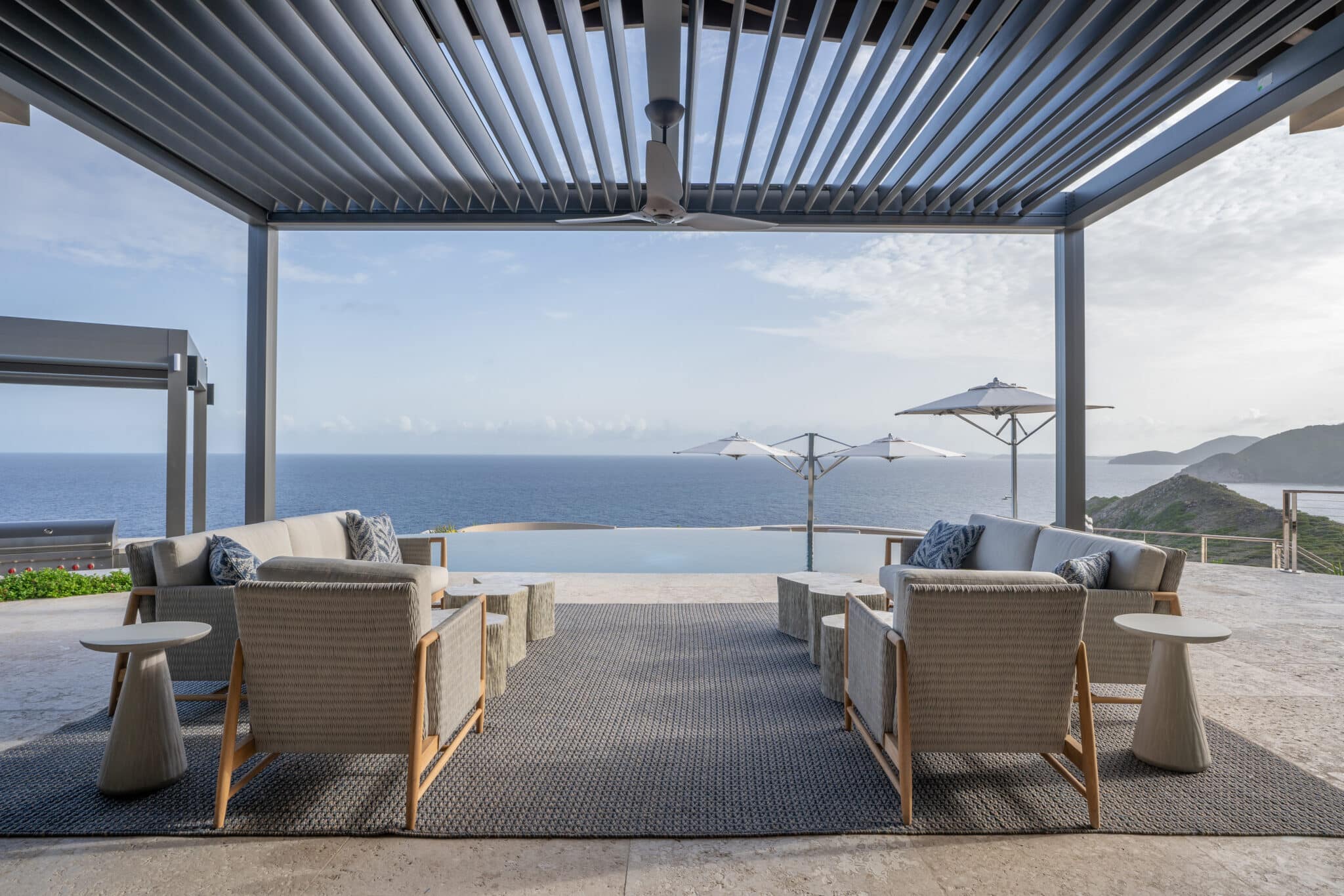 A spacious, modern outdoor terrace pavilion featuring an open-air louvered pergola roof, sleek grey wicker sofas and armchairs, and matching stone side tables facing a pristine infinity pool that blends seamlessly into the open ocean horizon.