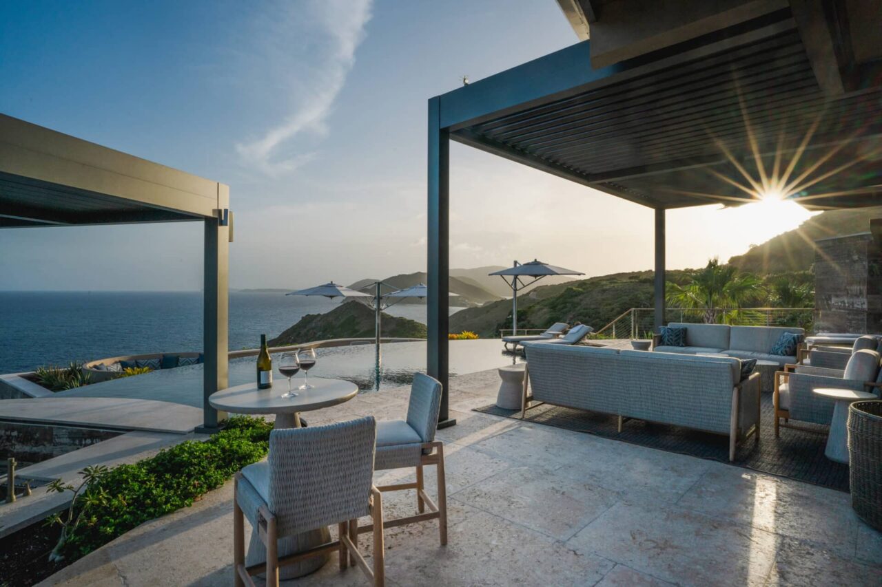 An expansive stone sun deck at Quintessa Villa featuring premium outdoor seating areas under a motorized louvered pergola next to a curving infinity pool, looking out over dramatic green rolling hills and a seaside cliff at sunset.