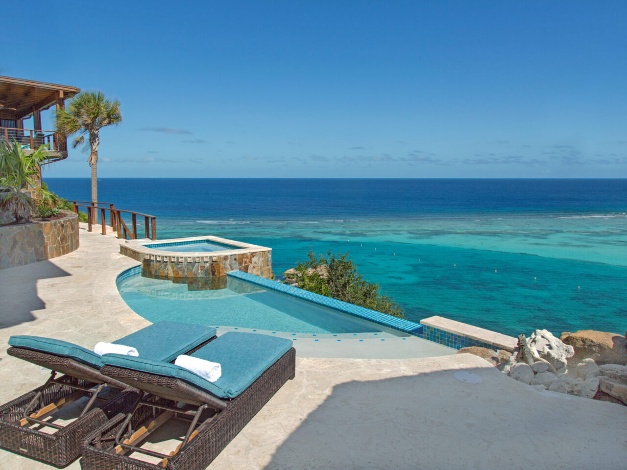Two cushioned blue lounge chairs side by side on a stone terrace overlooking a multi-level infinity pool and hot tub built with natural stone walls, framing a panoramic view of the turquoise reef-protected bay and deep blue ocean under a clear sky.