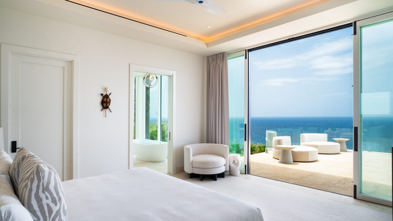 A bright, modern luxury bedroom at Oil Nut Bay featuring crisp white linens and a minimalist design. Large sliding glass doors open completely to a private stone terrace with white outdoor lounge chairs, showcasing an uninterrupted view of the deep blue Caribbean Sea under a clear sky.