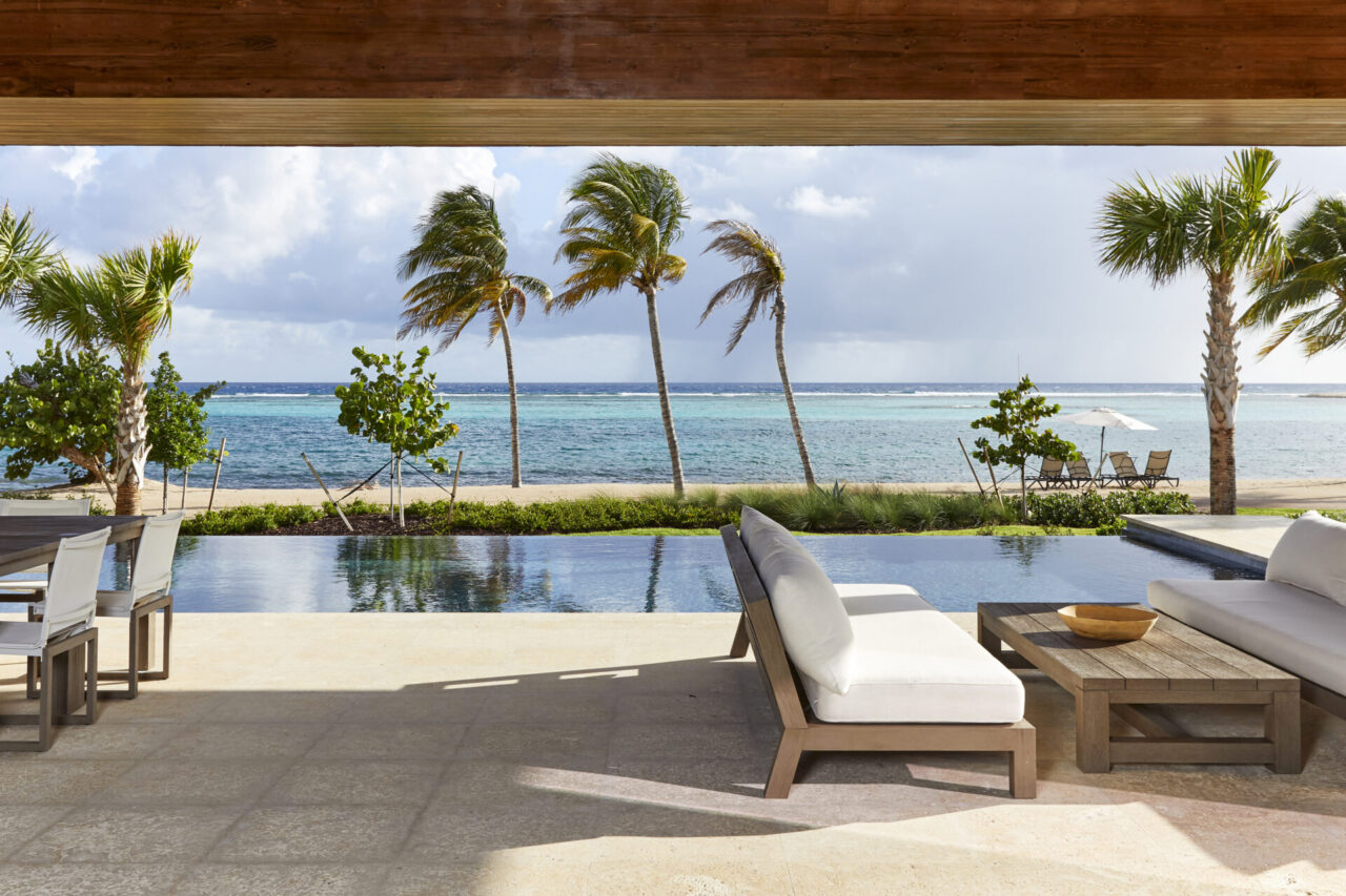View from the covered patio of The Beach House showing an outdoor lounge sofa and dining table next to a sleek infinity pool, with palm trees lining a pristine sandy beach and turquoise ocean waters in the background.