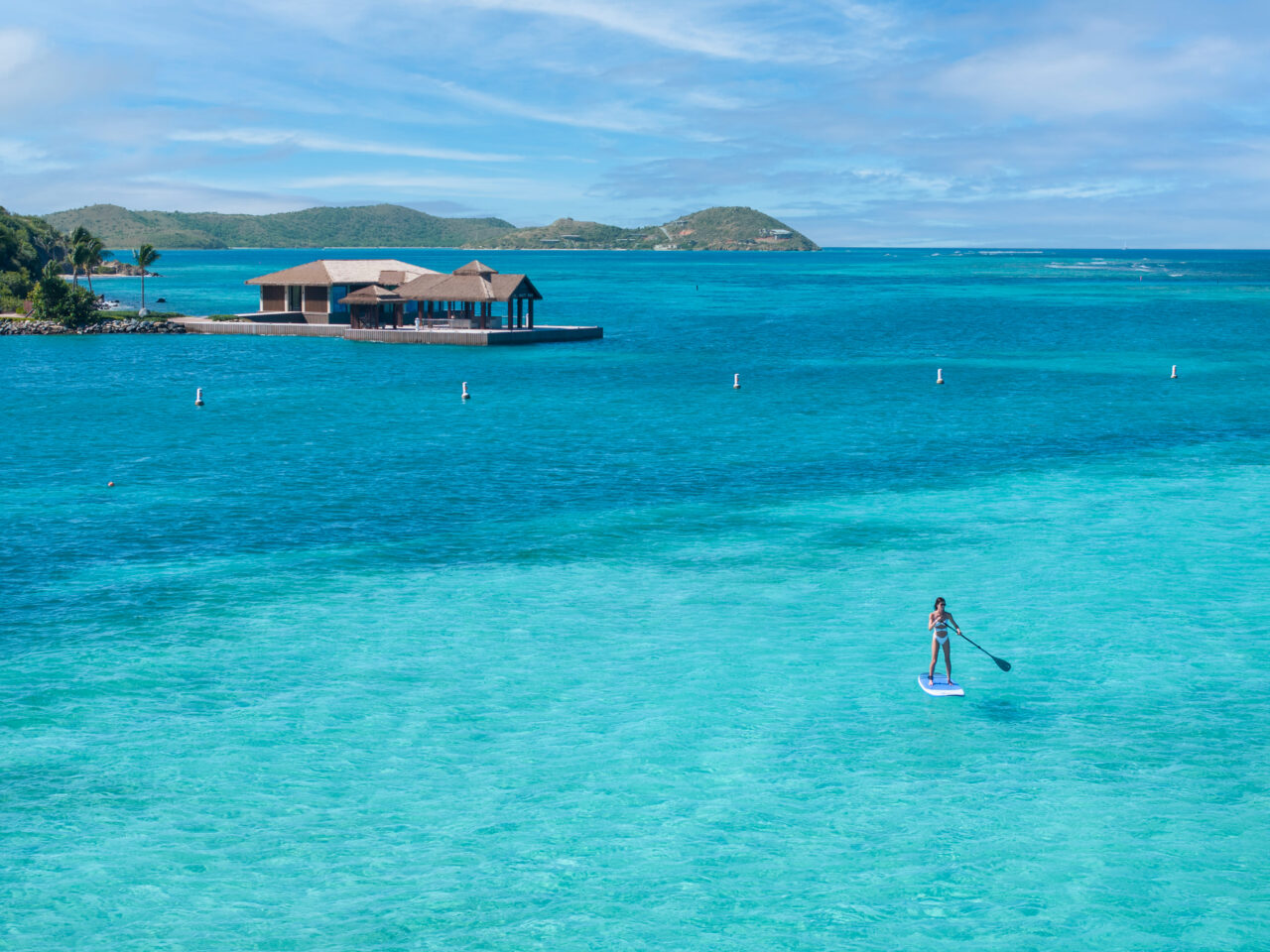 A scenic, eye-level view focusing on a woman stand-up paddleboarding in a vibrant tropical bay. Wearing a white bikini, she stands on a bright blue and white paddleboard, holding a paddle in the lower right foreground. The water transitions beautifully from a brilliant, shallow light turquoise near the paddleboarder to a deeper blue in the distance. In the background, a wooden overwater villa with thatched roofs sits on a platform near a green shoreline, with rolling green hills visible across the horizon under a lightly clouded blue sky.