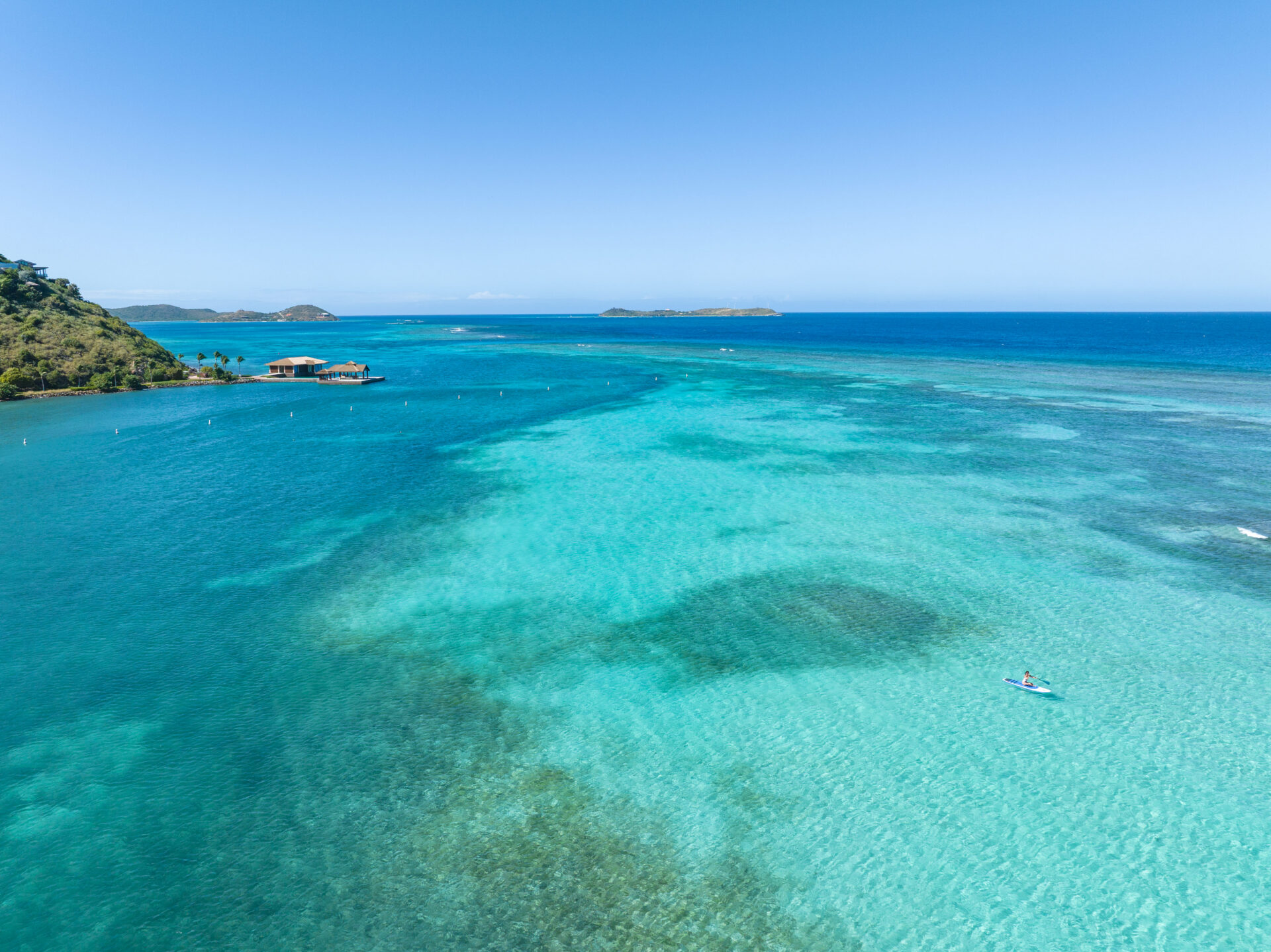 An aerial, high-angle view of a vast tropical bay with vibrant turquoise and deep blue water under a clear blue sky. In the lower right foreground, a single person is paddleboarding on a light blue board over a patch of clear, shallow water. In the distant left, a wooden overwater bungalow structure sits on a pier extending from a lush, green hillside coastline. Several small, green islands are visible on the horizon line where the bright sky meets the ocean.