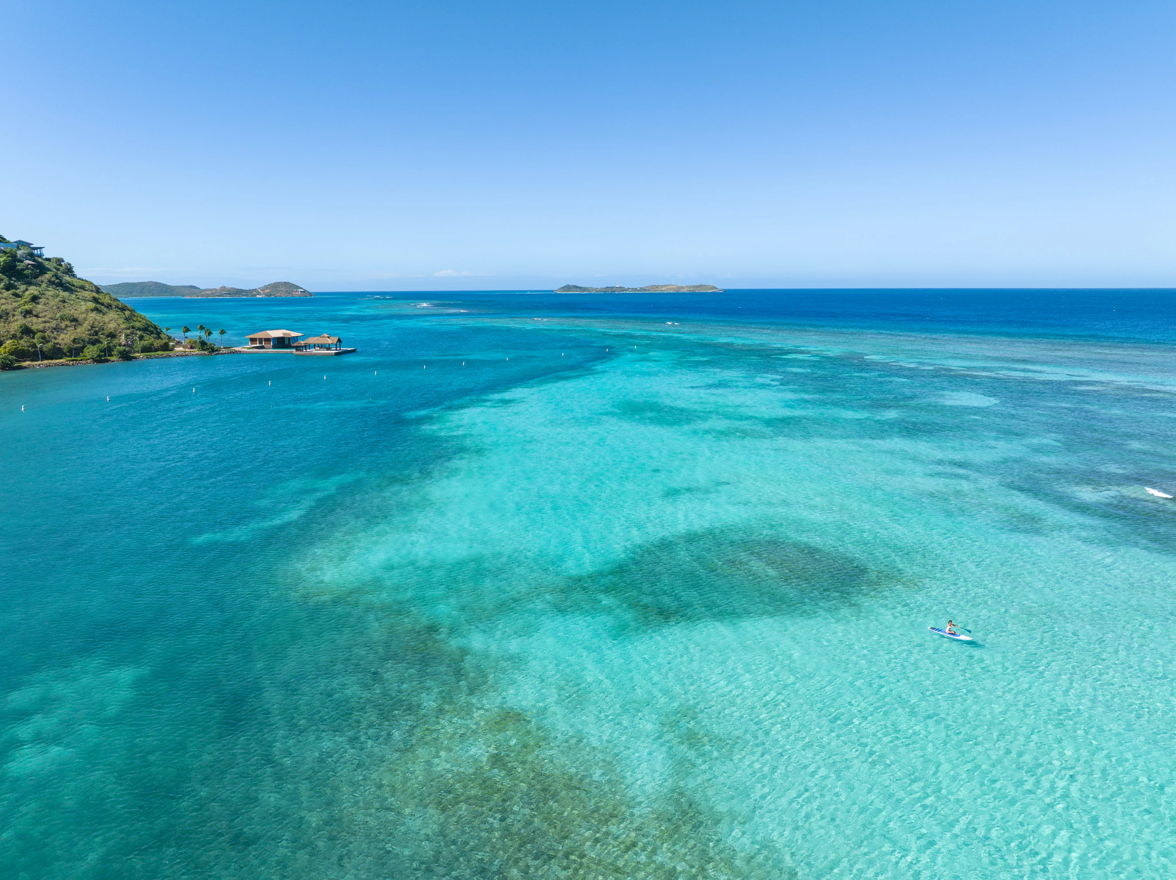 An aerial, high-angle view of a vast tropical bay with vibrant turquoise and deep blue water under a clear blue sky. In the lower right foreground, a single person is paddleboarding on a light blue board over a patch of clear, shallow water. In the distant left, a wooden overwater bungalow structure sits on a pier extending from a lush, green hillside coastline. Several small, green islands are visible on the horizon line where the bright sky meets the ocean.