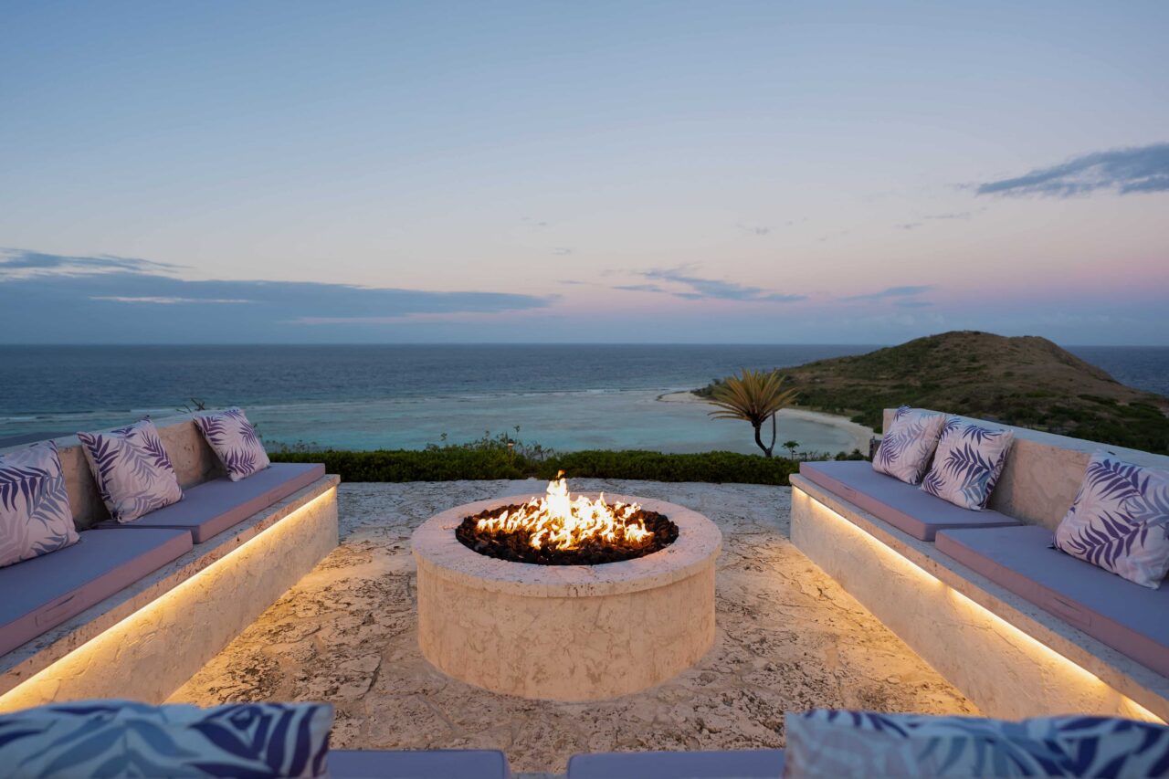A wide-angle, dusk shot of a luxurious outdoor stone patio at Oil Nut Bay, featuring a circular fire pit with active flames at the center. U-shaped stone benches with lavender-colored cushions and tropical-print pillows face the reef-protected bay and rolling green hills under a soft pink and blue evening sky.