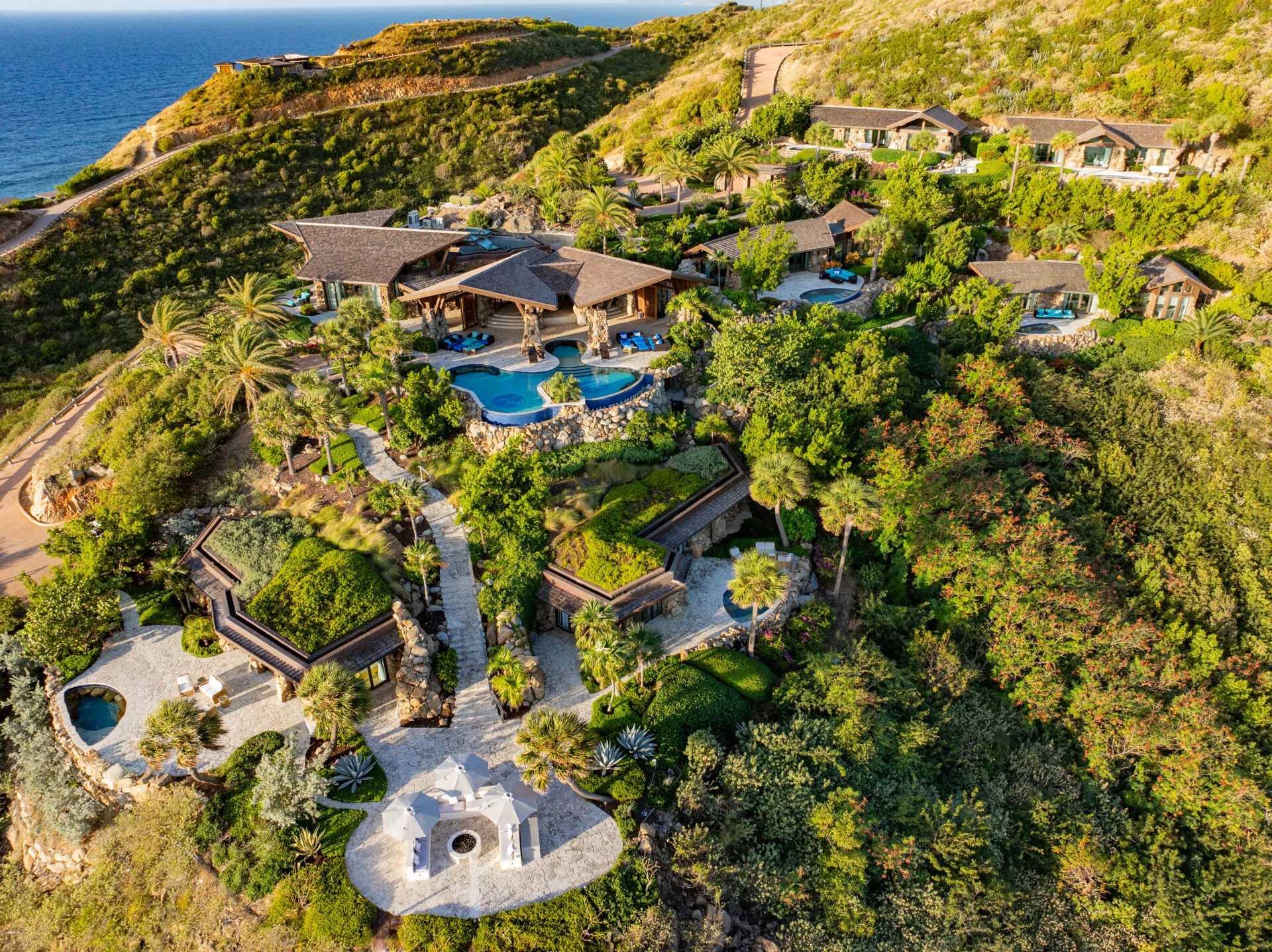 An aerial, high-angle view of a luxury hillside resort estate overlooking the deep blue ocean in the upper left corner. The sprawling property features multiple multi-level stone and wood villas with dark shingled roofs built directly into the lush, green tropical hillside. At the center of the estate sits a large, multi-tiered swimming pool with deep blue tiles, surrounded by palm trees, stone patios, and blue lounge chairs. Winding stone pathways and staircases connect the villas down to a lower circular patio area outfitted with white umbrellas, lounge seating, and a central outdoor fire pit. The entire compound is surrounded by dense green vegetation, trees, and steep hills under bright, warm sunlight.