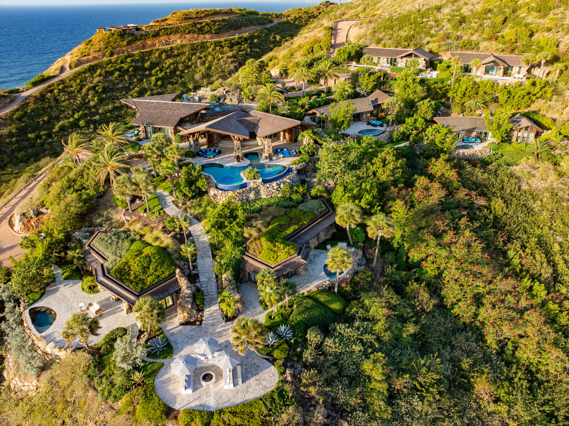 An aerial, high-angle view of a luxury hillside resort estate overlooking the deep blue ocean in the upper left corner. The sprawling property features multiple multi-level stone and wood villas with dark shingled roofs built directly into the lush, green tropical hillside. At the center of the estate sits a large, multi-tiered swimming pool with deep blue tiles, surrounded by palm trees, stone patios, and blue lounge chairs. Winding stone pathways and staircases connect the villas down to a lower circular patio area outfitted with white umbrellas, lounge seating, and a central outdoor fire pit. The entire compound is surrounded by dense green vegetation, trees, and steep hills under bright, warm sunlight.