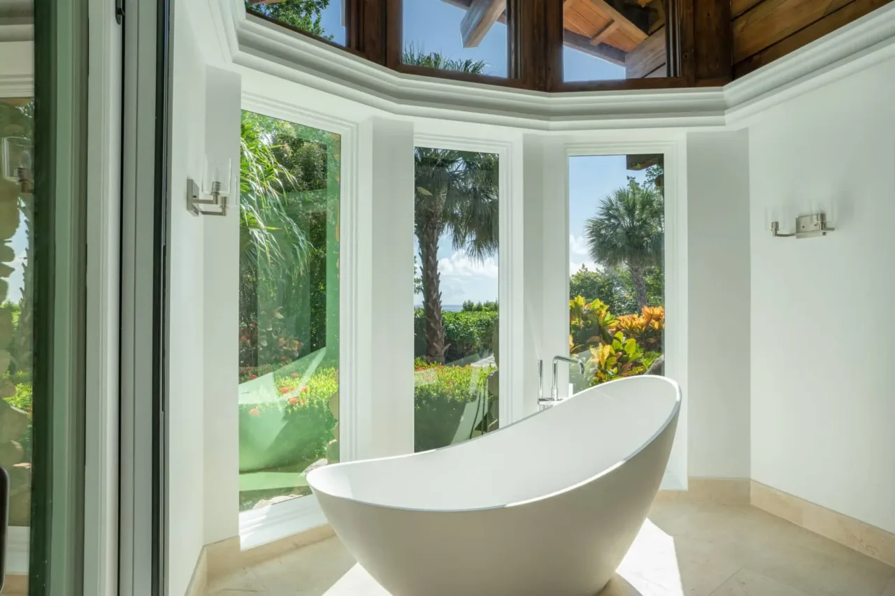 A modern white freestanding soaking tub positioned inside a sunlit, bay-window alcove of a luxury villa. Large vertical windows offer a pristine view of lush green tropical foliage, palm trees, and the ocean under a clear blue sky, set beneath an exposed dark wood ceiling frame.