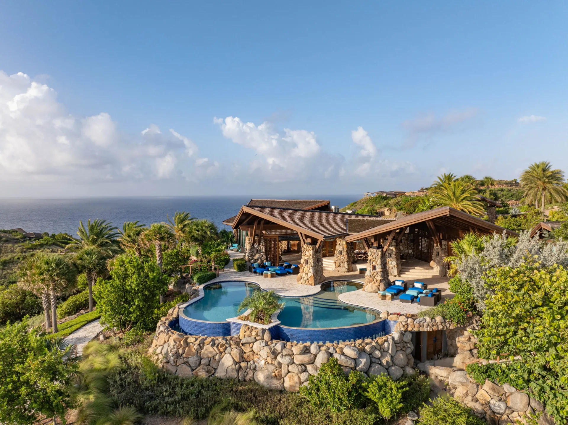 An aerial, high-angle view of a spectacular multi-level luxury estate nestled into the green cliffs of Virgin Gorda. The villa features distinctive natural stone pillars, dramatic exposed wood rooflines, a private multi-tiered infinity pool overlooking the deep blue Caribbean sea, and lounge decks surrounded by palm trees.