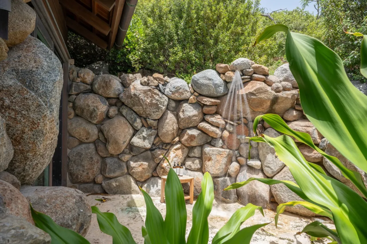 An outdoor luxury shower built seamlessly into a rustic natural stone wall at Oil Nut Bay. Lush green tropical foliage frames the foreground, and sunlight filters through the trees onto a small wooden side table holding premium bath products.