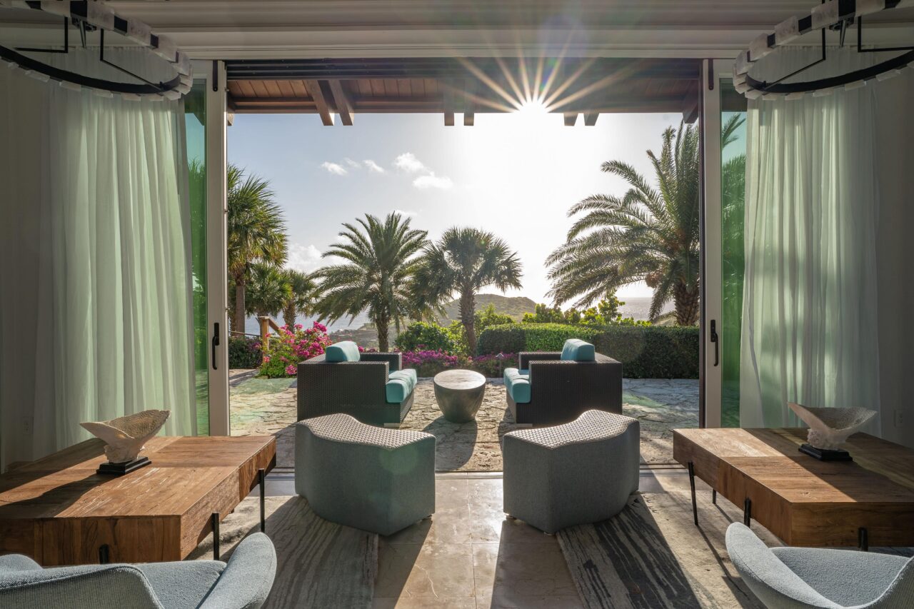 An interior view looking outward from a spacious, contemporary luxury living room through wide-open glass sliding doors. Sunbeams stream over palm trees and coastal greenery onto a stone patio with modern outdoor armchairs, while the foreground shows rustic wood coffee tables and chic, light blue lounge seating.
