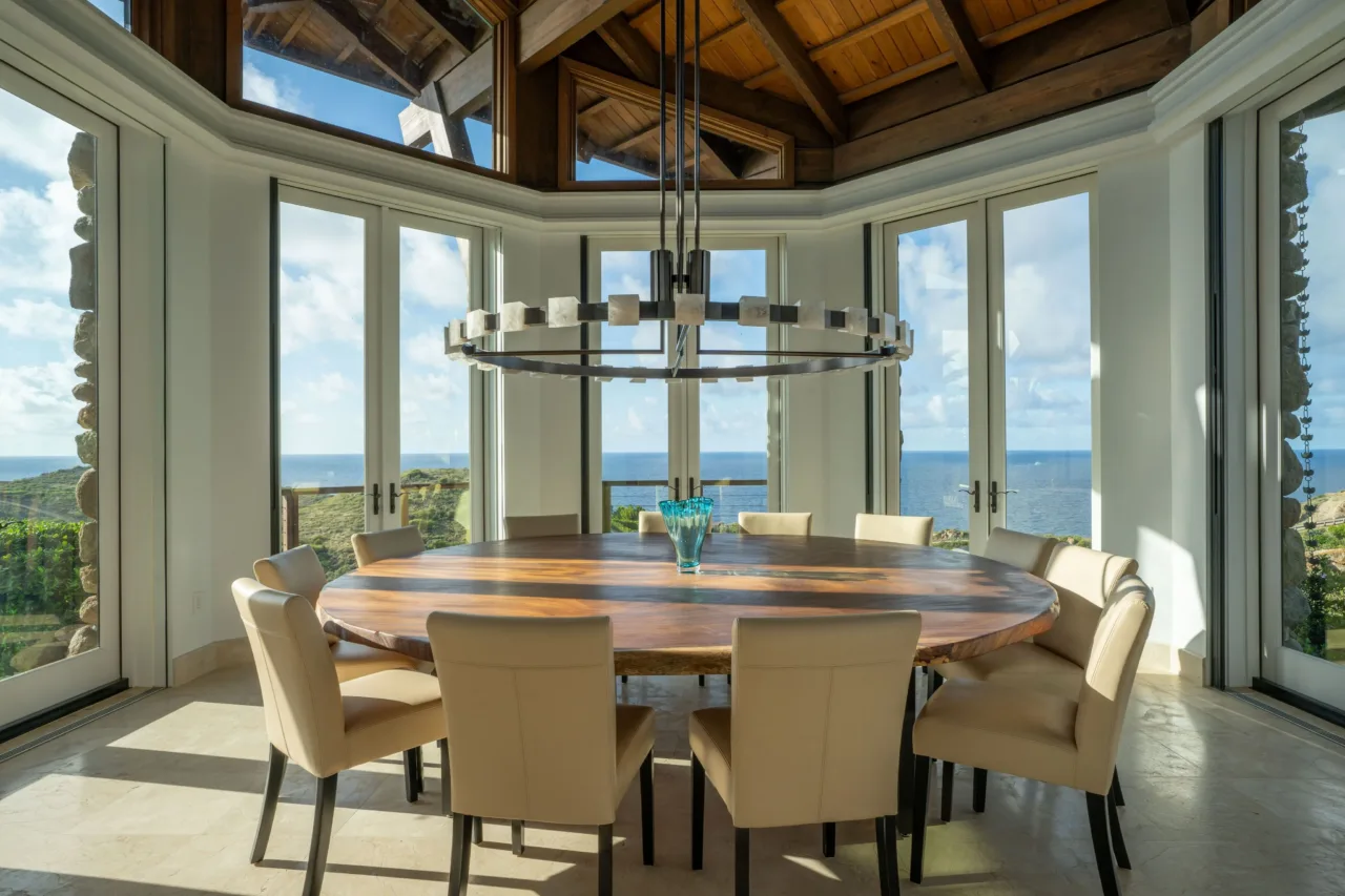 An elegant dining room with a large, polished round wood table and high-backed cream leather chairs under a dramatic modern ring chandelier. Expansive panoramic windows reveal a breathtaking view of green cliffs meeting the deep blue ocean.