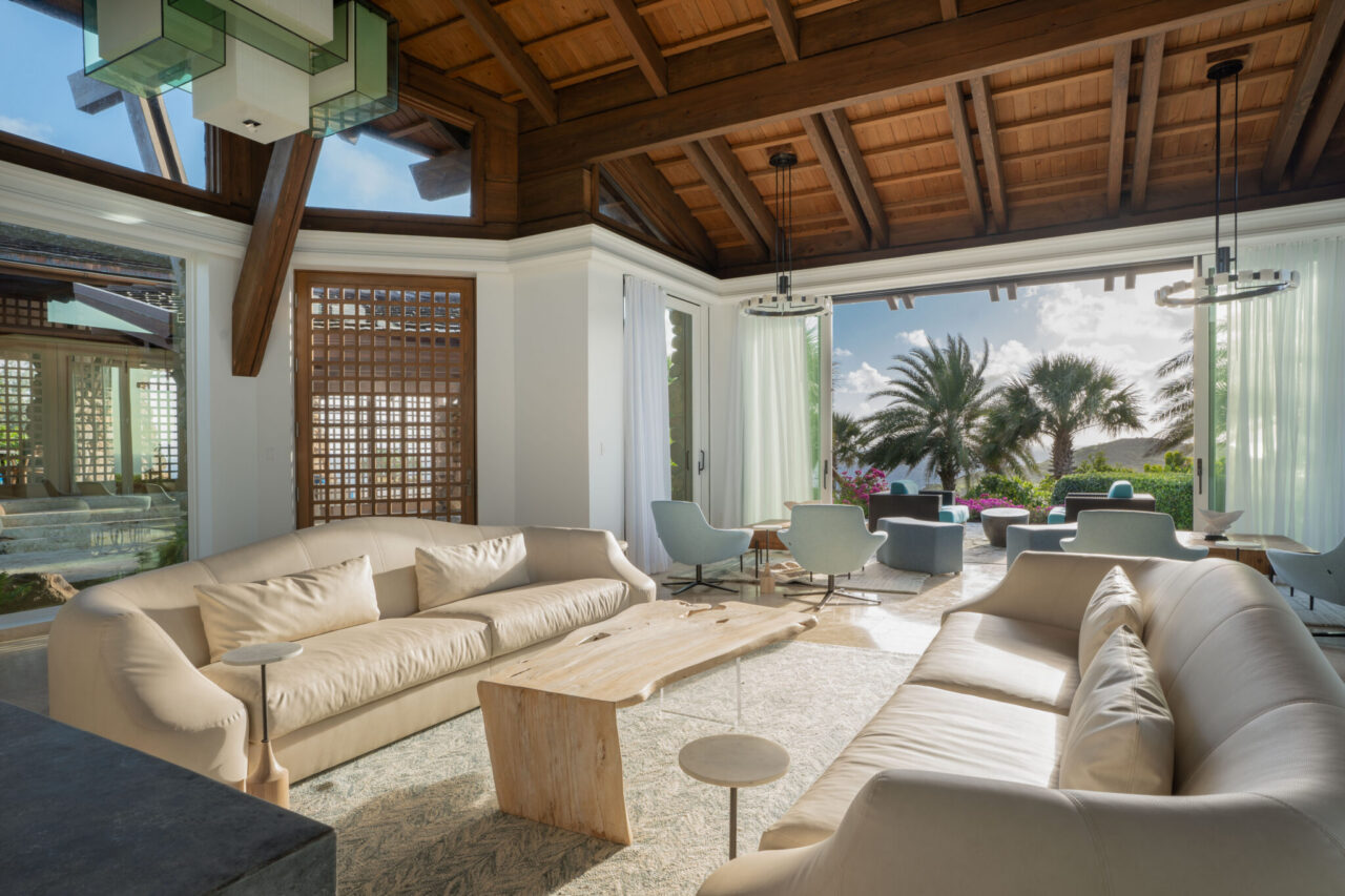 An elegant, open-concept villa living room showcasing organic architecture with high vaulted wood-beam ceilings, premium stone flooring, plush modern cream sofas, and a live-edge wooden coffee table looking out through massive floor-to-ceiling glass doors onto a tropical patio with palm trees and ocean views.