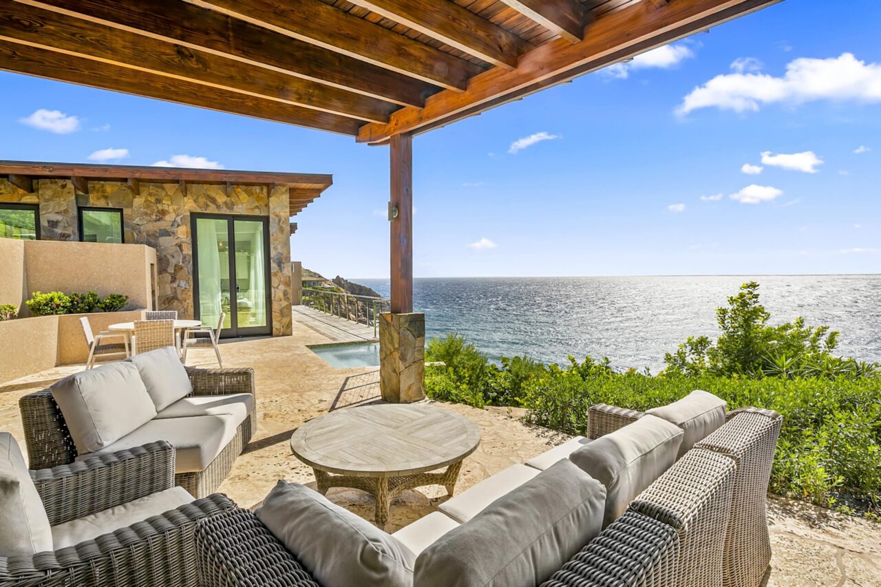 A covered stone patio at Sol Mate Villa furnished with comfortable woven outdoor sofas and a round wooden coffee table, framing a vast ocean view with the sun-drenched coastline extending into the sparkling sea under a blue sky.
