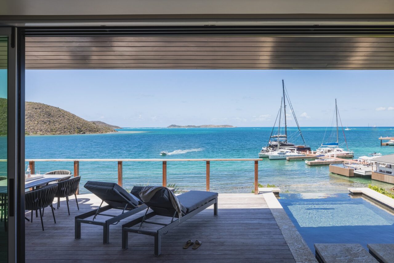 View from a luxury villa terrace at Oil Nut Bay, showcasing premium sun loungers, an infinity plunge pool, and an al fresco dining table overlooking sailing yachts docked at the reef-protected North Sound.