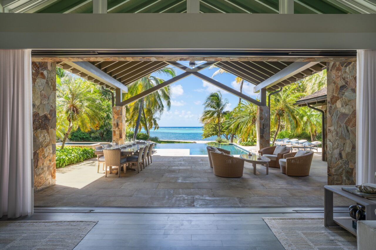 A view looking out from a spacious stone-walled great room through expansive pocket doors onto a covered pavilion terrace featuring an outdoor dining table, lounge seating, a private pool, and the turquoise Caribbean Sea framed by palm trees.