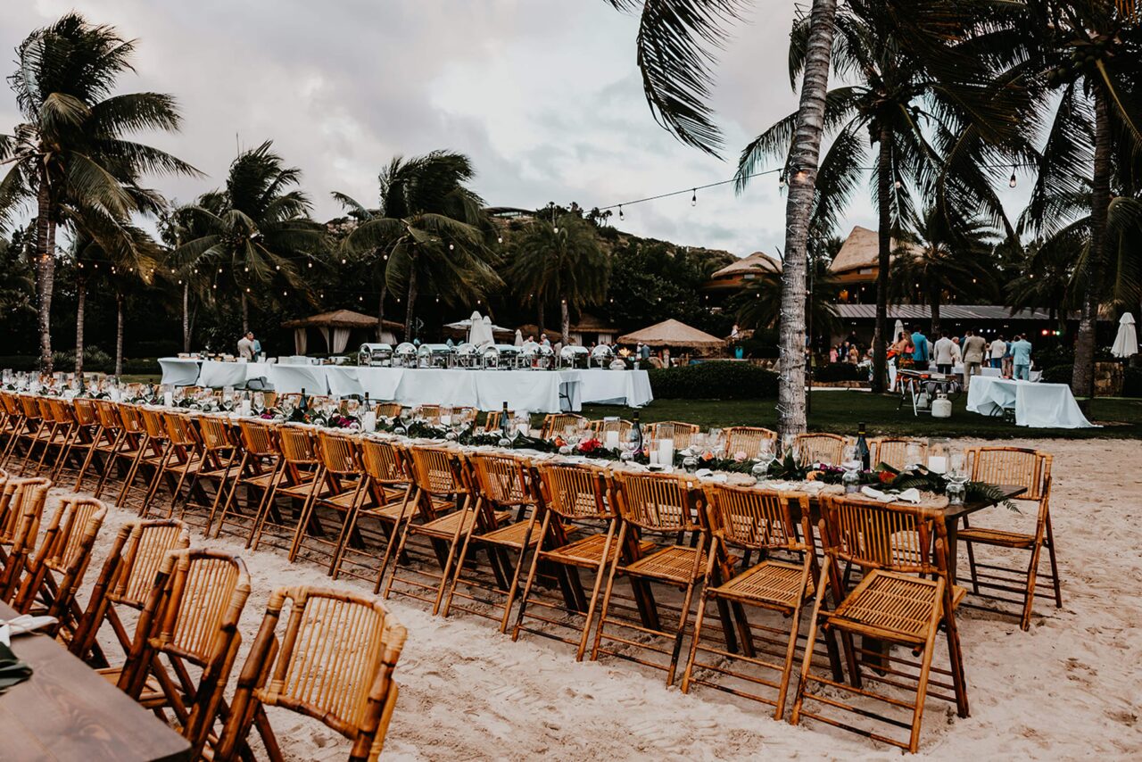 An elegant beachfront wedding reception dinner setup on the sand at Oil Nut Bay, featuring long wooden tables, bamboo chairs, green garlands, and illuminated palm trees at dusk.