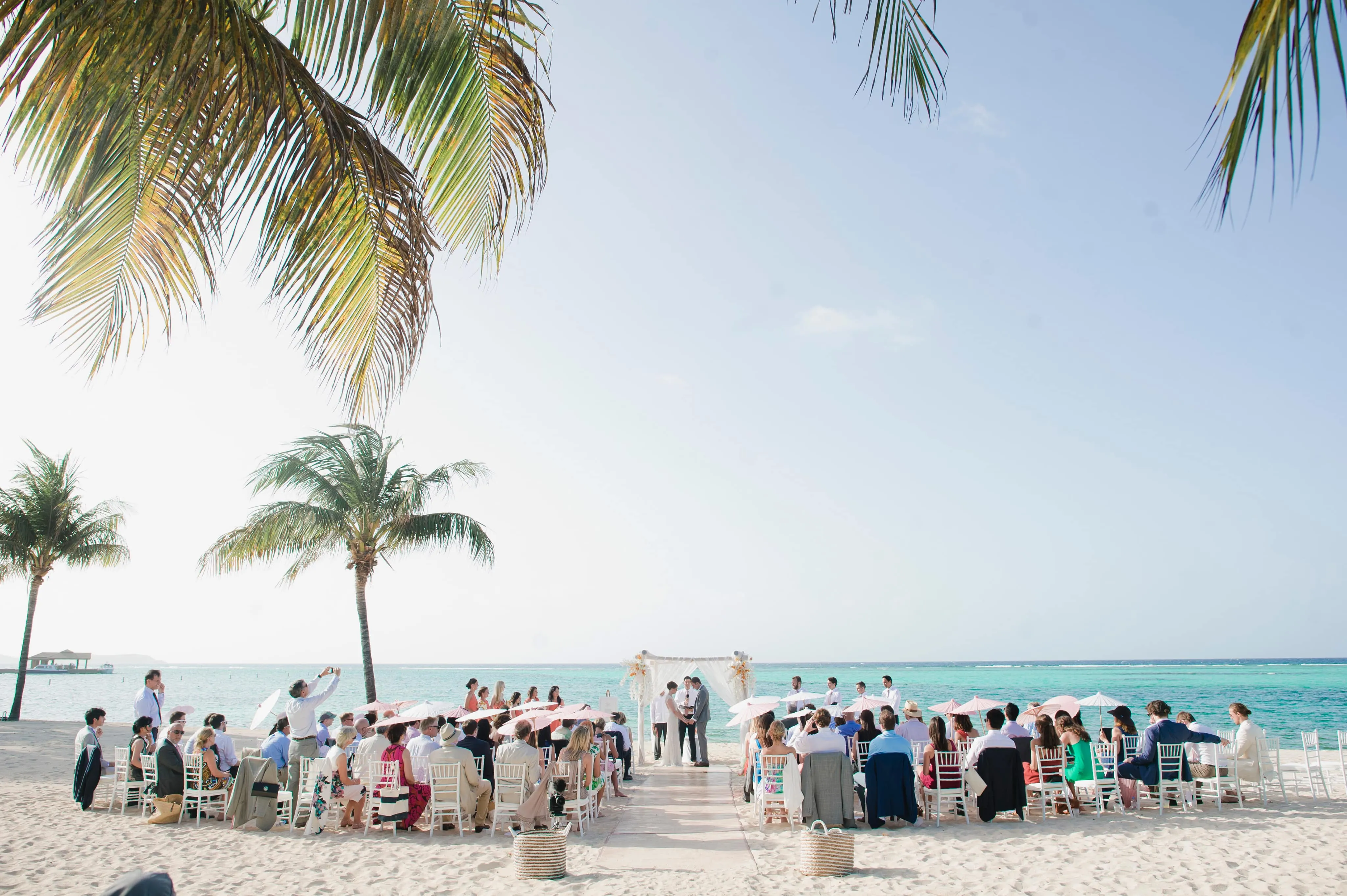 A pristine beach wedding ceremony at Oil Nut Bay with guests seated in white chairs holding pink parasols, facing a white floral arch overlooking the turquoise Caribbean Sea.