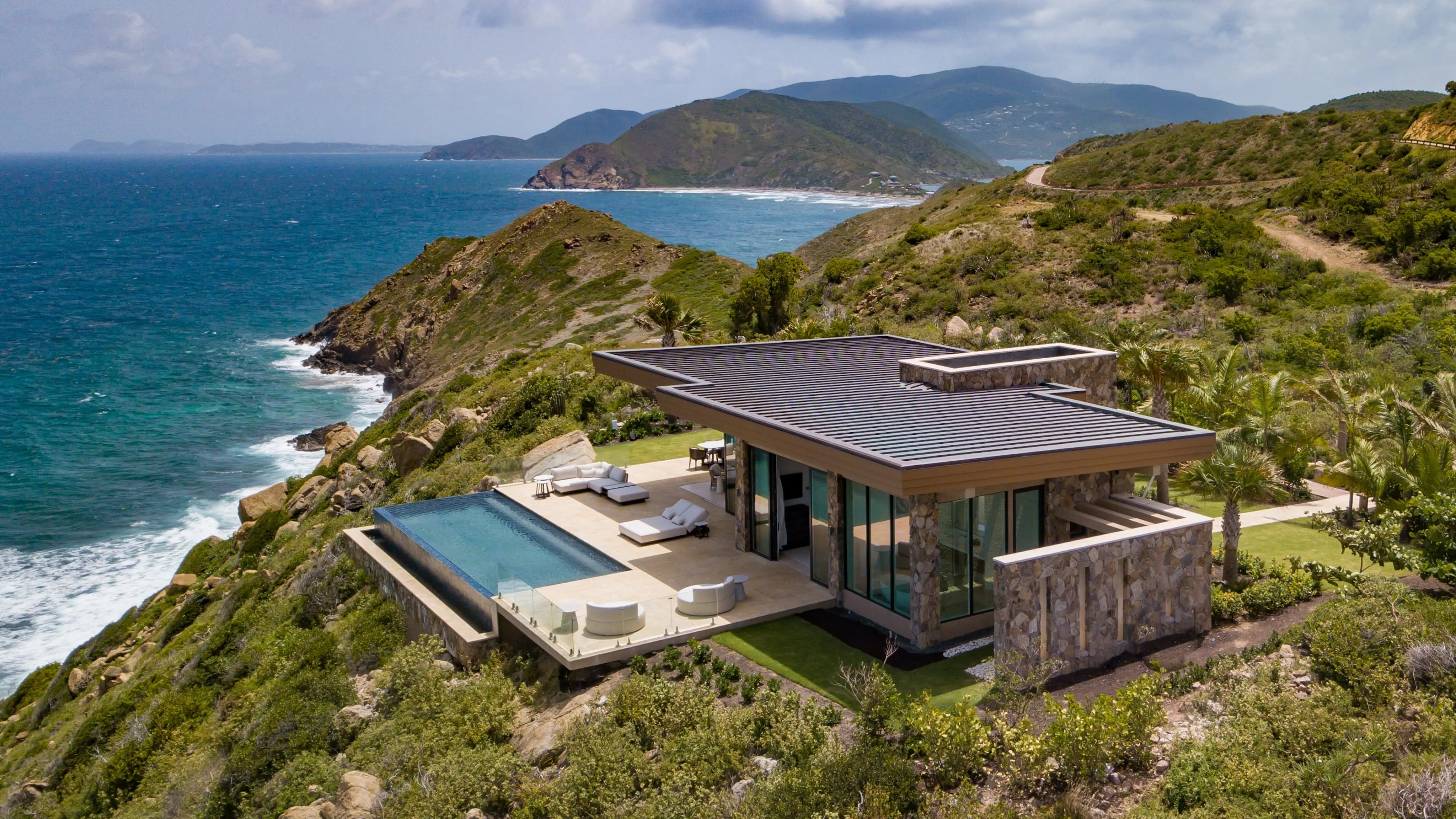 An aerial drone photograph of a sleek, modern luxury villa built into a steep green hillside on a cliff edge, featuring a private infinity pool, sun loungers, and expansive views of the British Virgin Islands coastline.