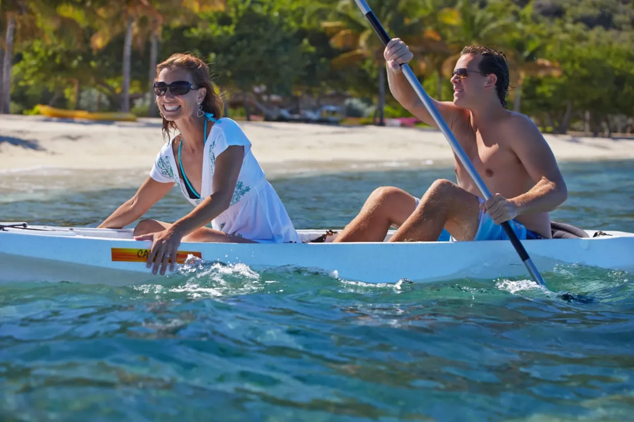 A smiling couple paddling a white tandem kayak through the clear, turquoise reef-protected waters along the white-sand beach at Oil Nut Bay in the British Virgin Islands.