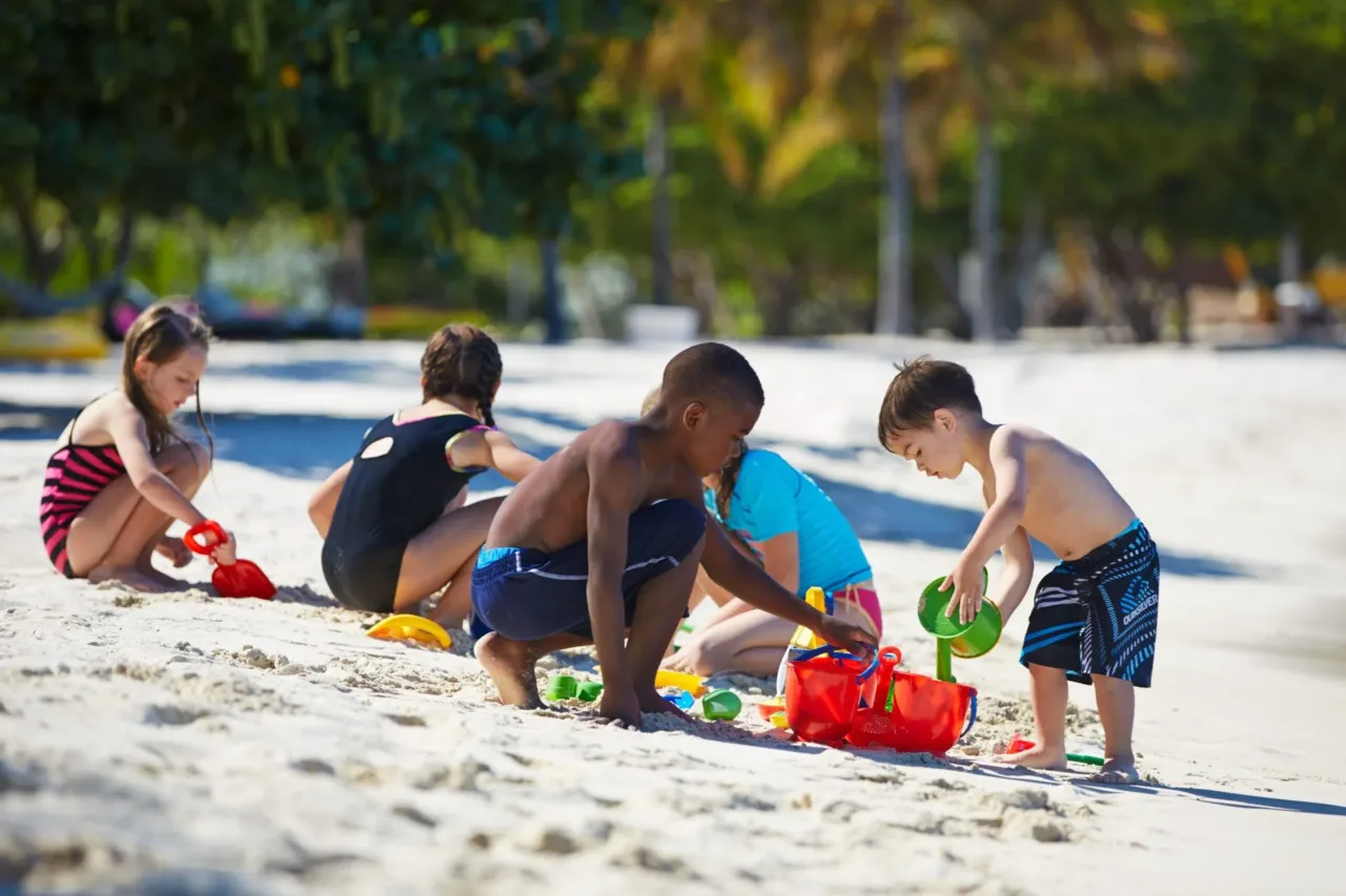 A group of young children playing safely with colorful buckets and shovels in the soft white sand beach under the shade of trees, demonstrating luxury family nanny services at Oil Nut Bay resort.