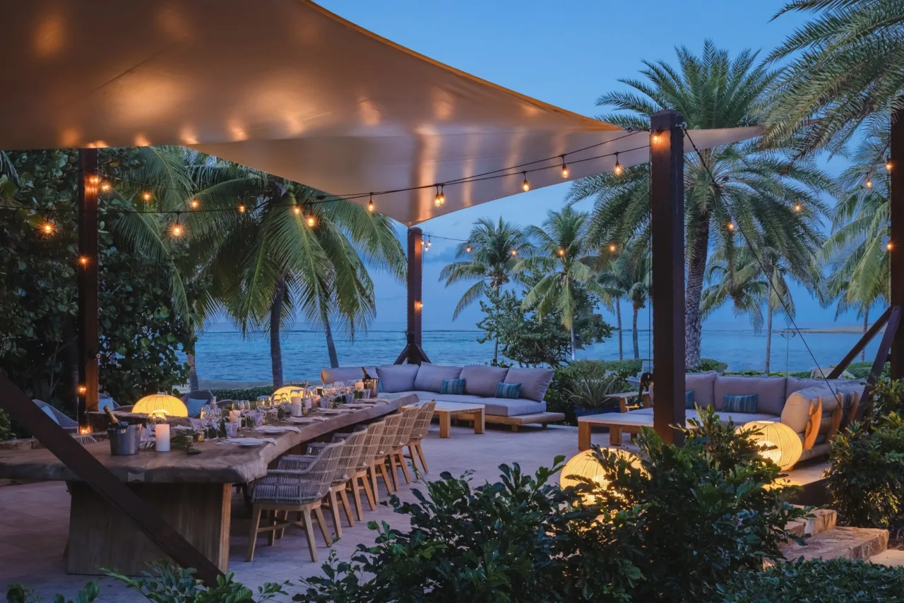 An intimate outdoor dining setup at the Beach Club Marlin Deck in Oil Nut Bay at twilight, featuring a long live-edge wooden table under a canvas canopy, glowing string lights, chic woven chairs, surrounding lounge sofas, and palm trees overlooking the calm Caribbean Sea.