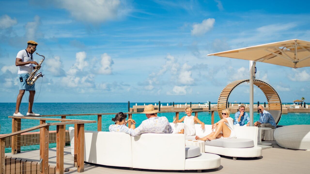 Guests relaxing on white contemporary outdoor sofas on an oceanfront deck at Oil Nut Bay’s Marina Village while a live musician plays the saxophone under a clear blue sky.