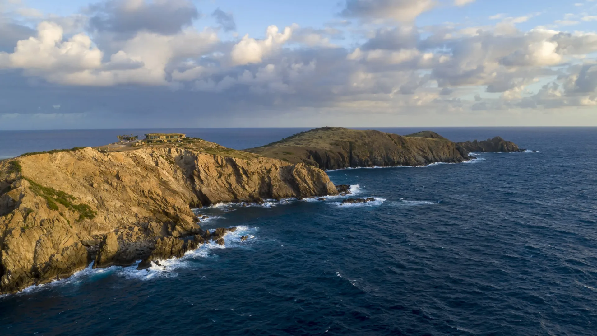 A wide aerial drone shot of a jagged, rocky peninsula cliffside meeting the churning deep blue sea under a cloudy sky, with a secluded estate nestled on the highest ridge point.