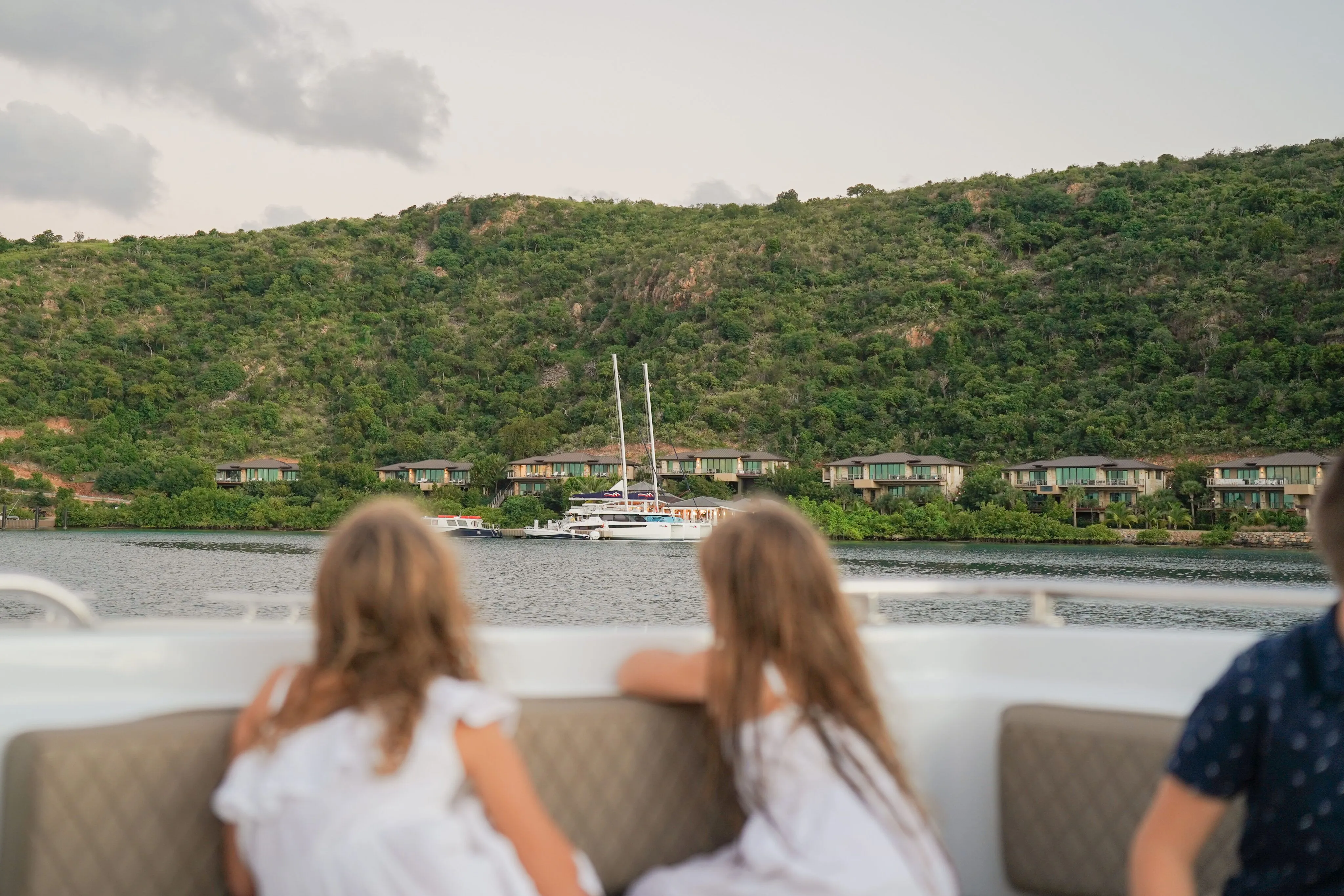 Two young girls sitting at the bow of a luxury boat charter, looking toward the exclusive waterfront properties and marina slips of Oil Nut Bay nestled at the base of a dramatic, verdant tropical ridgeline.