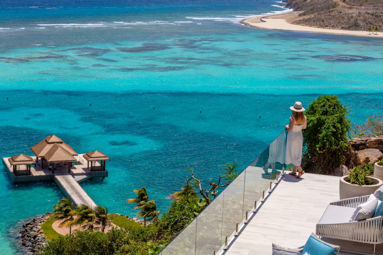An elevated, high-angle shot captures a woman in a white sundress and straw hat standing on a modern stone terrace with a glass railing, looking out over a vibrant, turquoise tropical bay. Below, a wooden walkway leads to a floating dock with three thatched-roof gazebos, while the clear blue ocean reveals a shallow coral reef stretching toward a curved, sandy beach and a dry, hilly coastline in the distance.