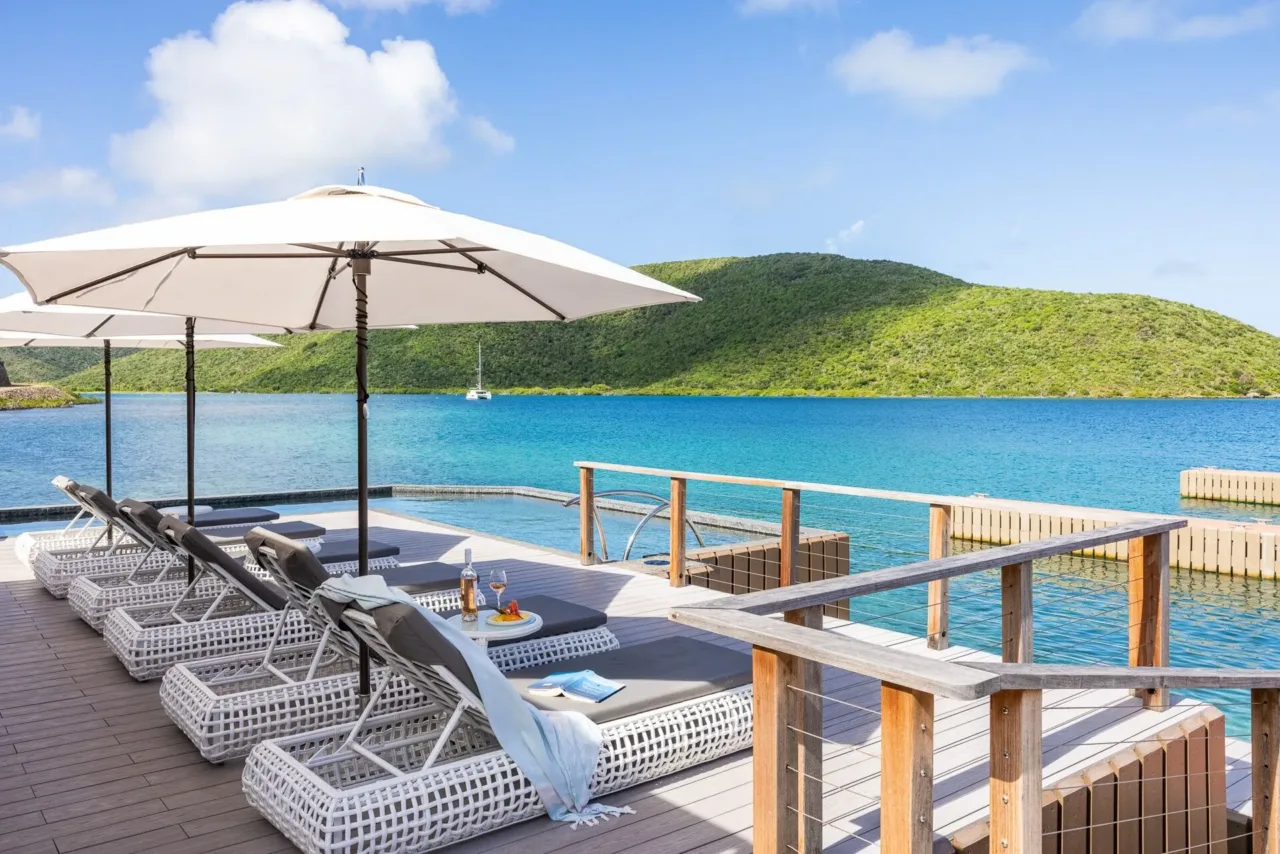 Elegant white wicker sun loungers lined up on a pristine wooden dock terrace at Oil Nut Bay Marina Village, looking out over calm deep-water channels toward lush green tropical hillsides on Virgin Gorda.