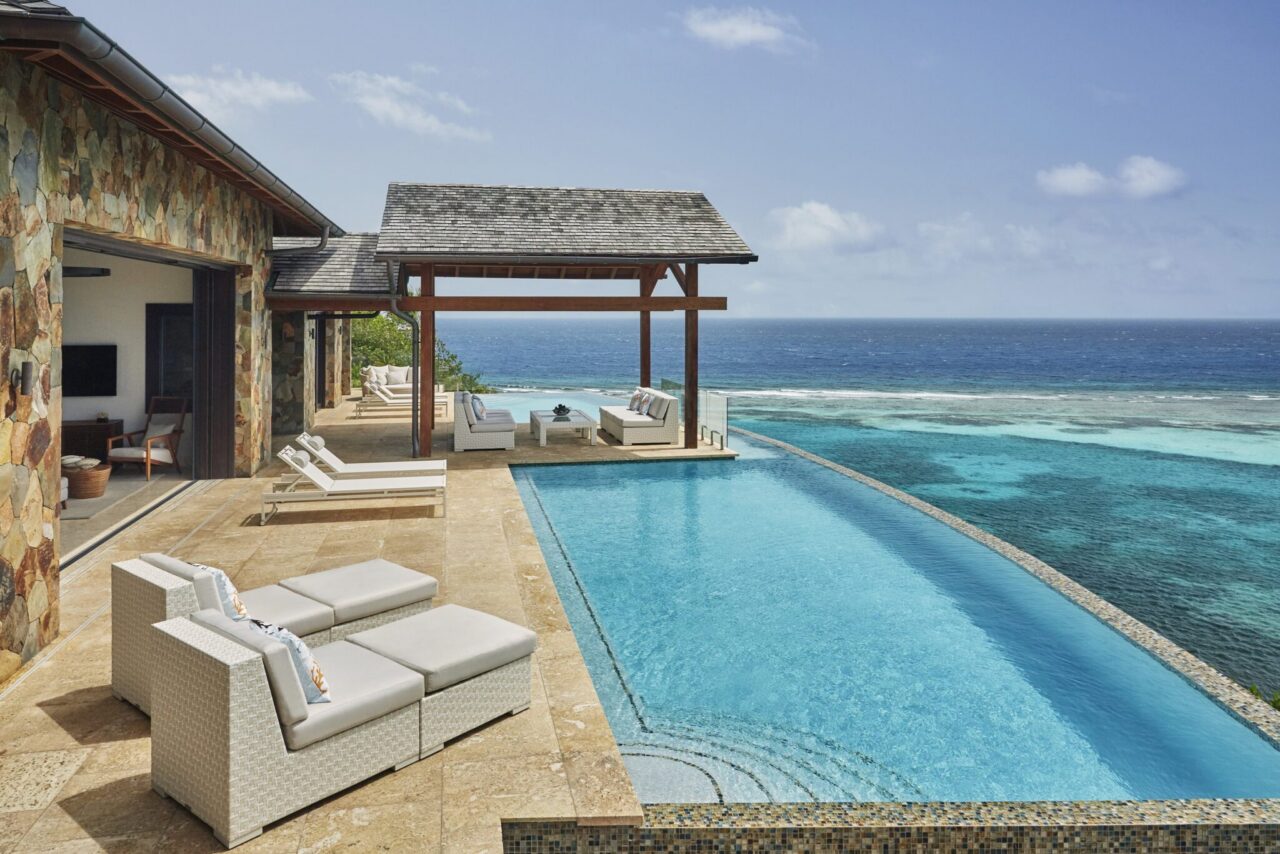 Elevated view of a private infinity-edge swimming pool flanking a stone-clad luxury villa terrace with plush white sun loungers and a shaded wooden gazebo overlooking a vibrant coral reef-protected bay.