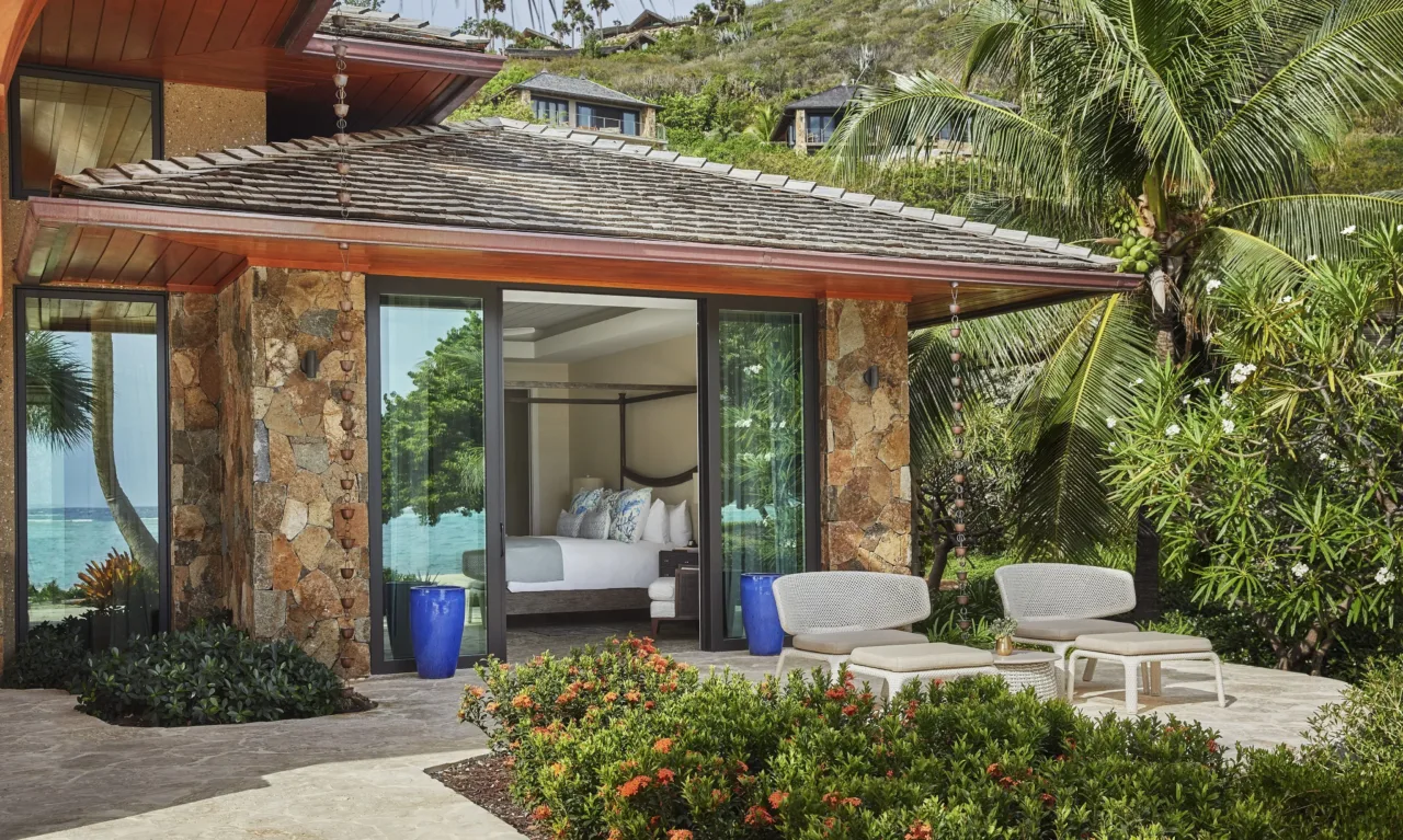 Exterior view of a luxurious natural stone beach villa pavilion with a wooden shingle roof, showing a bedroom with a four-poster bed through open glass sliding doors next to an outdoor stone patio with lounge chairs amidst lush tropical landscaping.