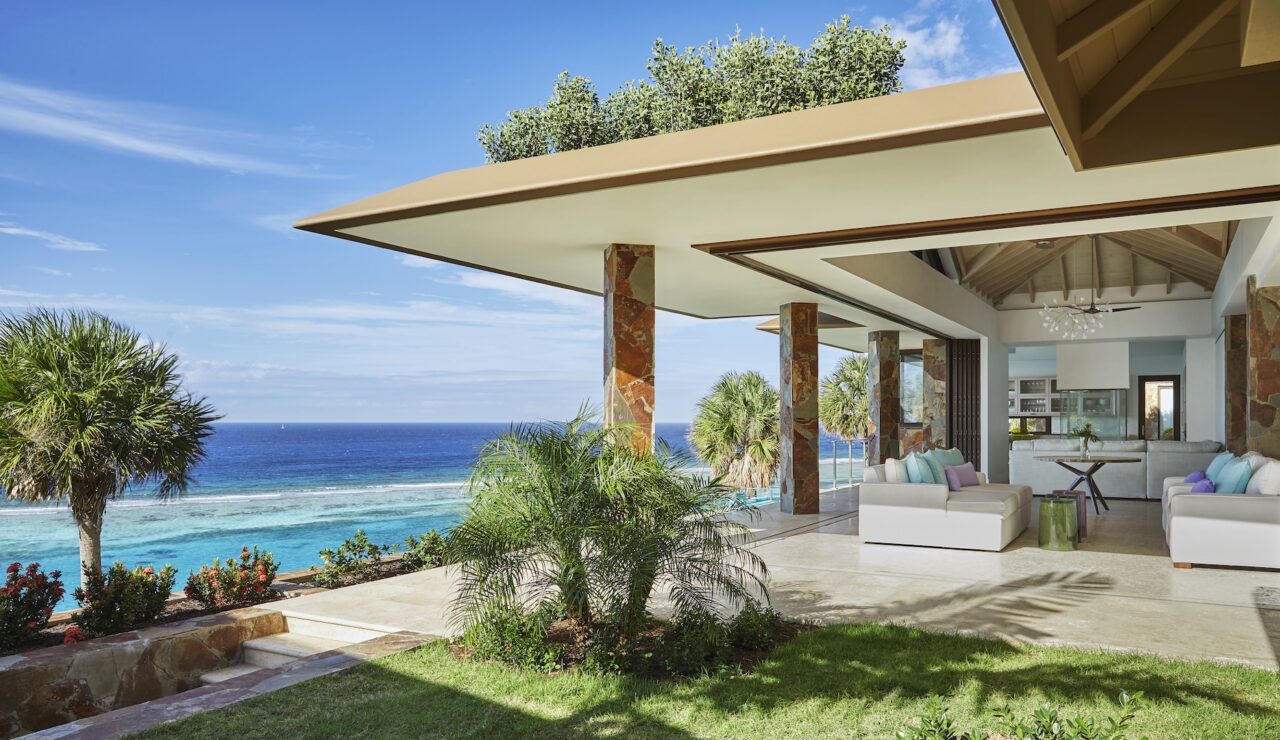 Covered stone-pillared veranda of Villa Montastraea at Oil Nut Bay, featuring luxury white lounges with pastel accents overlooking a coral reef and the deep blue Caribbean Sea.