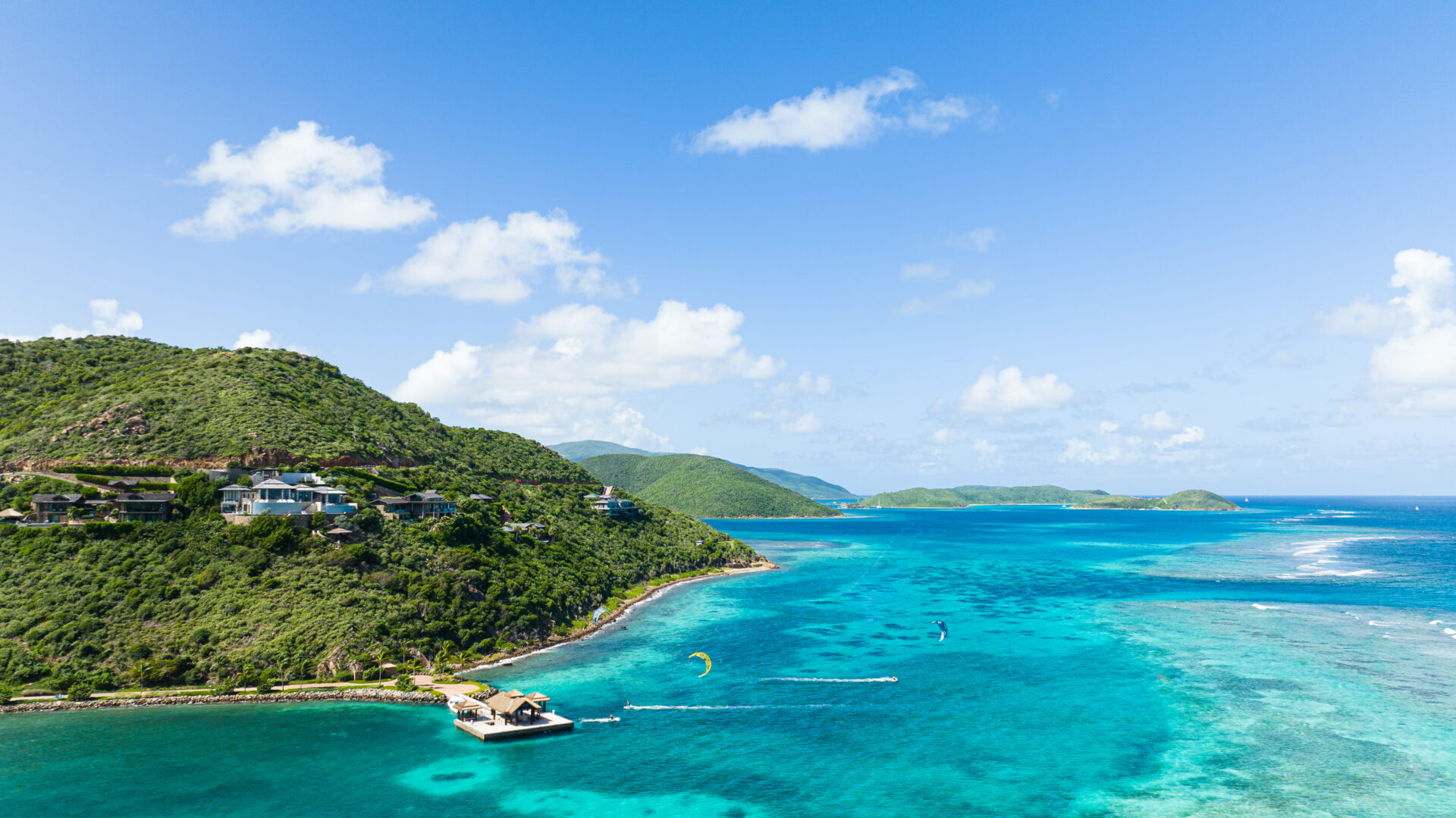 High aerial drone view of the lush green peninsula at Oil Nut Bay, showing the private overwater arrival dock, hillside luxury real estate, and kiteboarders on the bright turquoise reef waters.