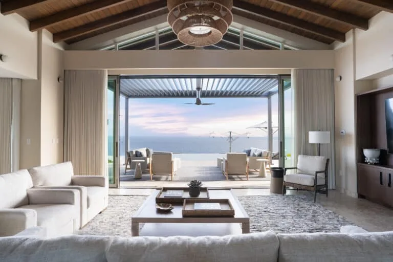 Spacious, modern open-air living room inside a luxury estate at Oil Nut Bay, featuring high vaulted wooden ceilings, contemporary white sofas, and massive sliding glass doors opening to a covered patio overlooking the ocean at sunset.