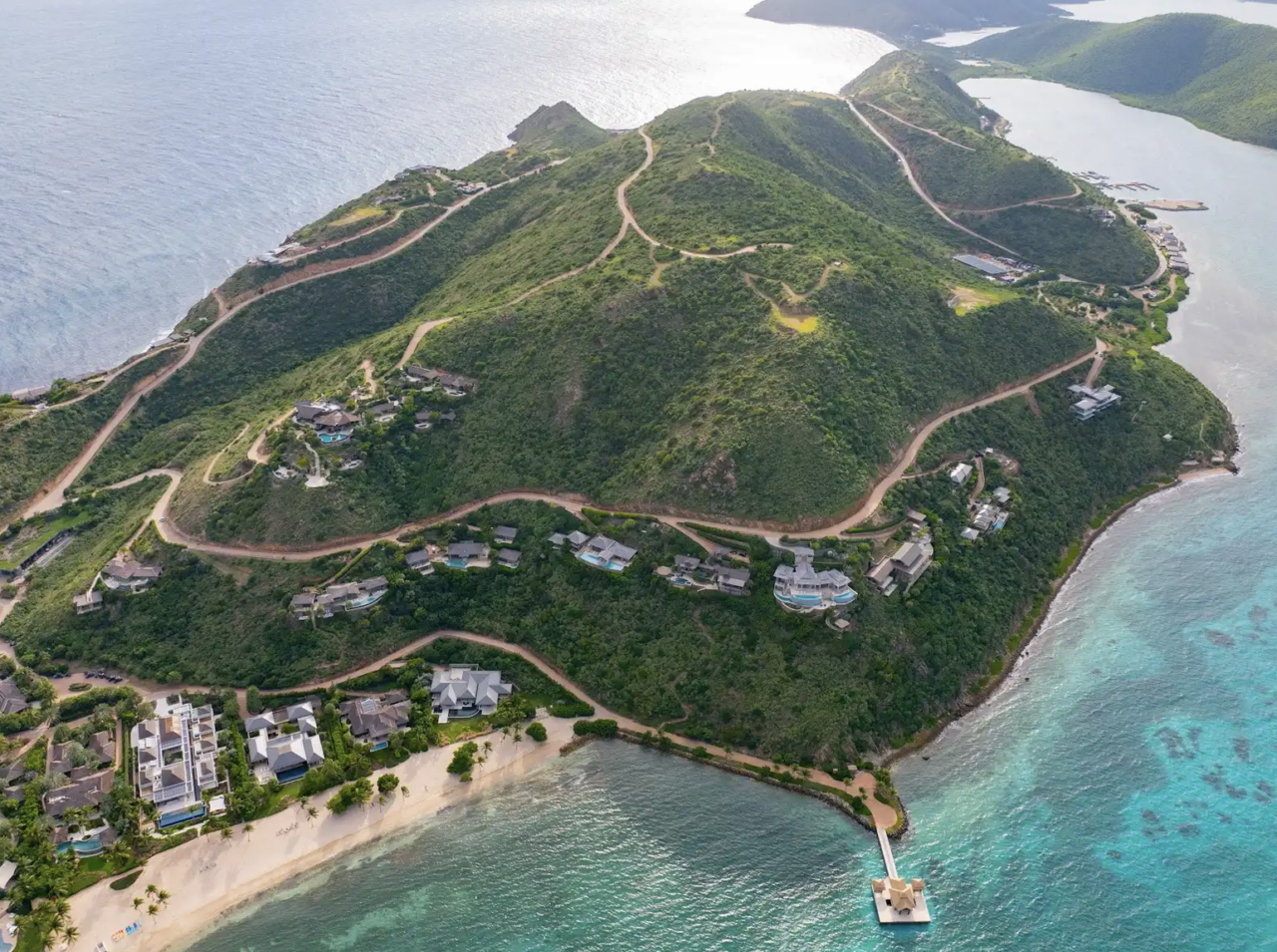 An aerial, high-angle view shows a lush, green peninsula covered in winding dirt roads and luxury hillside villas overlooking the ocean. At the bottom right, a wooden walkway leads out to a floating dock with thatched-roof gazebos, situated in clear, light-turquoise water. The left side features a sandy beach lined with beachfront properties and palm trees, while the steep hills of the peninsula rise dramatically toward a ridge line overlooking the deeper blue sea.