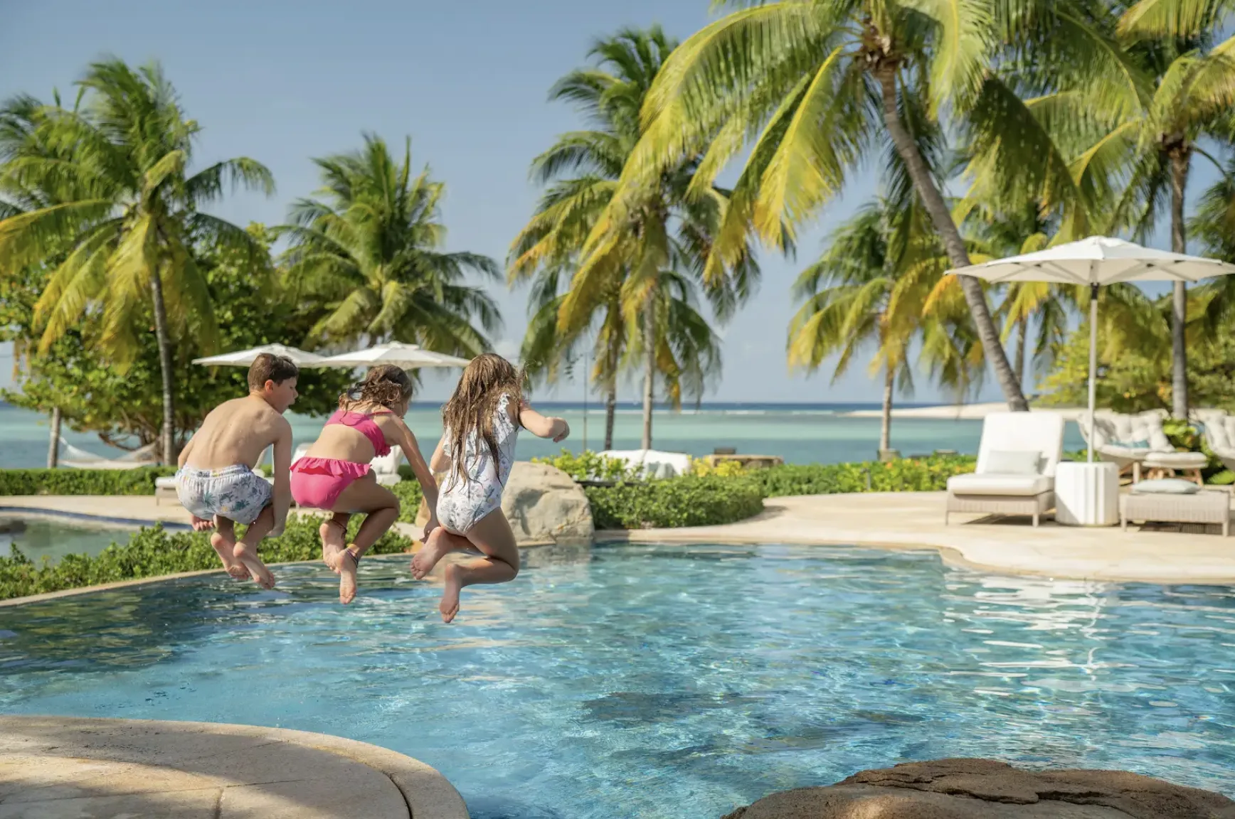 Three children are captured mid-air as they joyfully jump together into a clear, blue swimming pool at a tropical resort. The pool area is surrounded by lush green foliage and tall palm trees, with white lounge chairs and patio umbrellas set up along the stone deck. In the background, the calm turquoise ocean meets a clear sky under bright, sunny daylight.