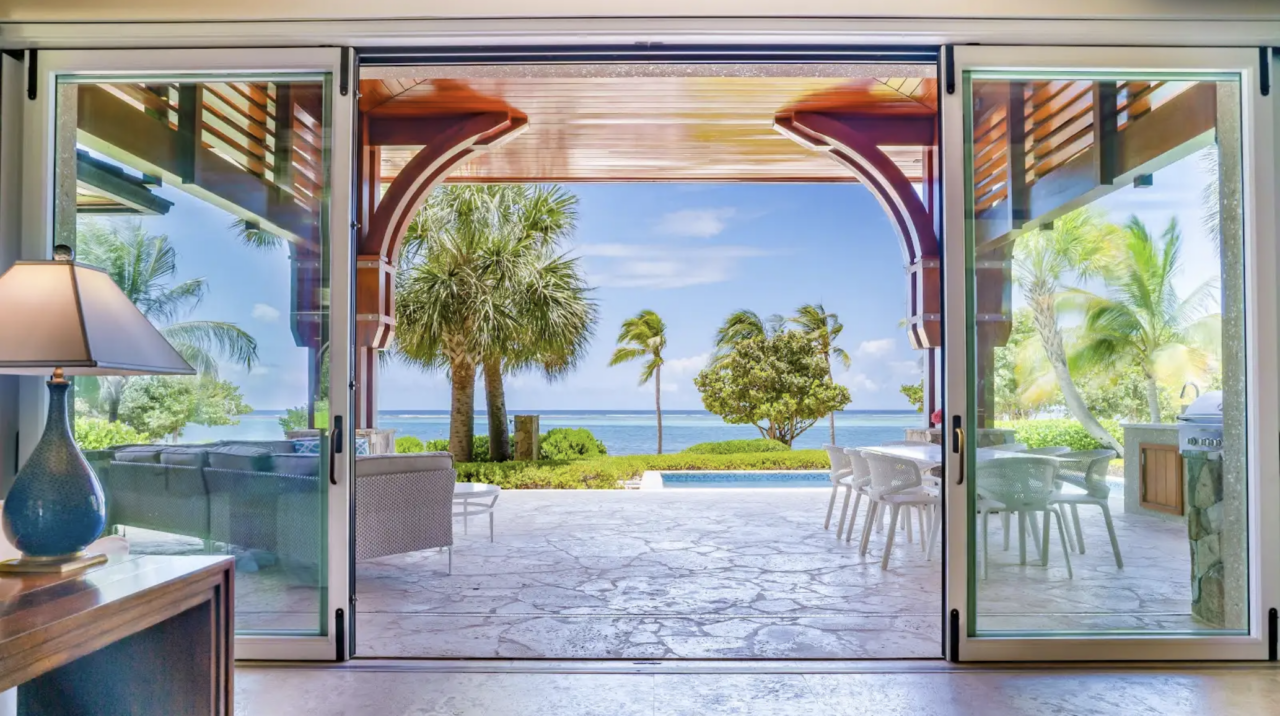 A wide shot framed by open, sliding glass doors reveals a luxurious covered patio with a polished wooden ceiling and stone-tiled flooring. The patio features a comfortable gray sectional sofa on the left and a long white outdoor dining table with an adjacent kitchen area on the right. Beyond the terrace, a manicured green lawn with palm trees and a small pool leads directly to a view of the calm, blue ocean under a bright sky.