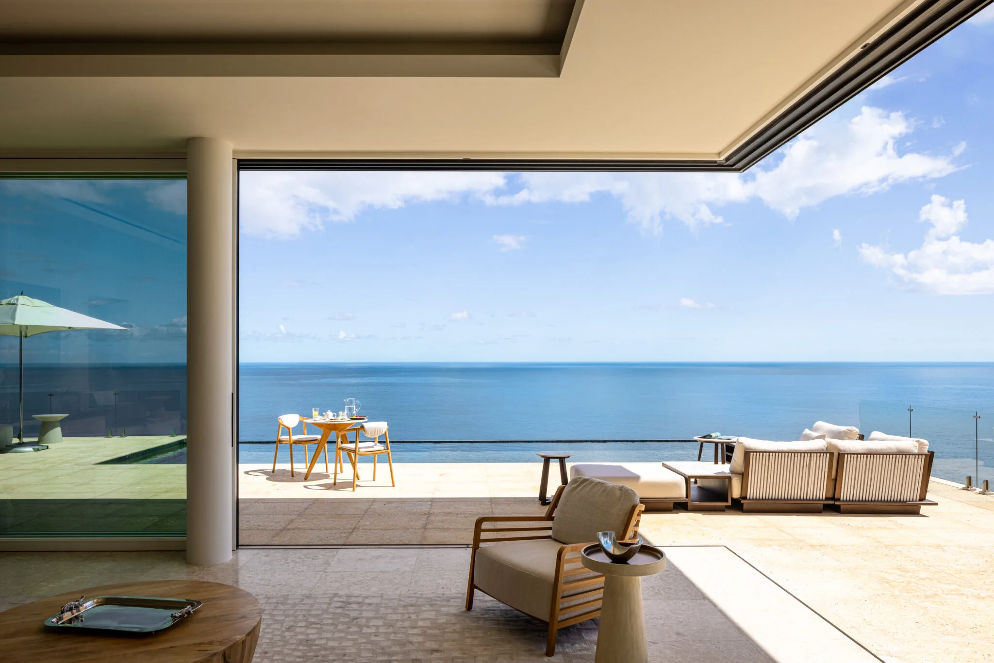 High-angle interior perspective of a modern minimalist villa lounge opening completely via corner glass doors to a sun-drenched stone terrace with an outdoor dining table, lounge seating, and an endless view of the deep blue Atlantic horizon.