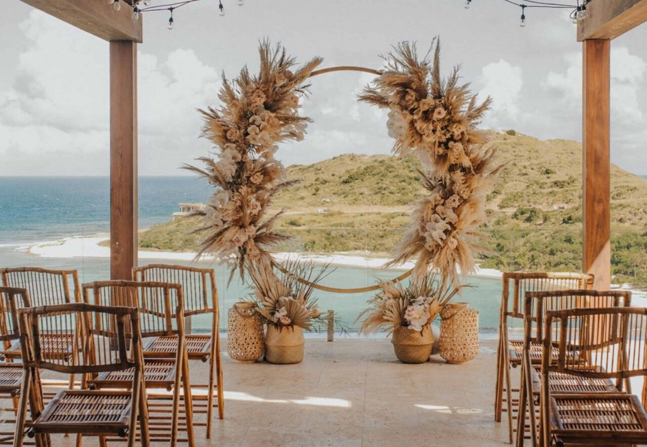 A luxury outdoor wedding ceremony venue on a covered stone terrace at Oil Nut Bay, featuring a circular golden arch decorated with bohemian pampas grass and soft blush roses, bamboo folding chairs, and panoramic views of the turquoise bay and tropical hills.
